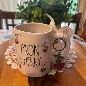 May include: A cream-colored ceramic mug with the words "MON CHERRY" in black script, adorned with red cherry illustrations. A cherry charm hangs from the handle. The mug sits on a wooden tray with a white lace doily.