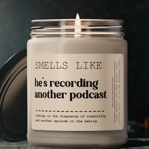 May include: A white candle in a glass jar with a black lid. The label reads "SMELLS LIKE he's recording another podcast" and "Indulge in the fragrance of creativity and another episode in the making."