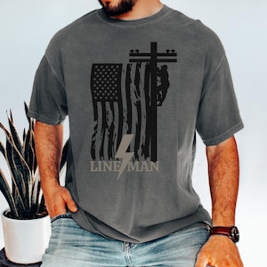 May include: A gray t-shirt with a black and white graphic of a lineman working on a power pole with an American flag in the background. The text "LINE MAN" is printed below the graphic.