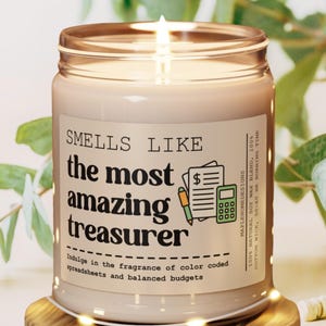 May include: A beige candle with a brown lid and a white label that reads "Smells Like the most amazing treasurer". The label also includes a green and yellow illustration of a calculator, a pen, and a piece of paper with a dollar sign. The text on the label reads "Indulge in the fragrance of color coded spreadsheets and balanced budgets".