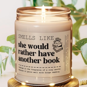 May include: A lit candle in a glass jar with a label that reads "Smells Like she would rather have another book." The label also features a graphic of books and a teacup. The candle is on a wooden coaster.