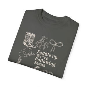 May include: Dark gray t-shirt featuring white line drawings of cowboy boots, a cowboy on a horse, a lasso, a cowboy hat, and a cactus. The text "Saddle Up We're Following Jesus" is also printed on the shirt.