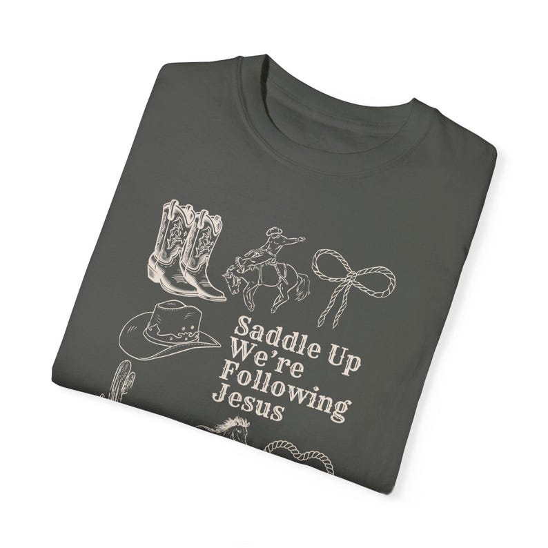 May include: Dark gray t-shirt featuring white line drawings of cowboy boots, a cowboy on a horse, a lasso, a cowboy hat, and a cactus. The text "Saddle Up We're Following Jesus" is also printed on the shirt.