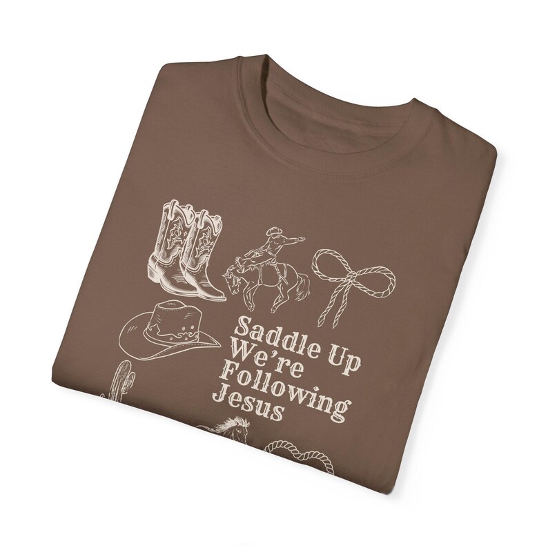 May include: A brown t-shirt featuring a white graphic of cowboy boots, a cowboy on a horse, a lasso, a cowboy hat, and the text "Saddle Up We're Following Jesus."