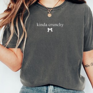 Op de afbeelding: Een grijs T-shirt met de tekst "kinda crunchy" en een witte strik bedrukt op de voorkant.
