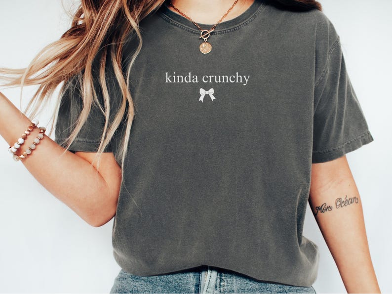 Op de afbeelding: Een grijs T-shirt met de tekst "kinda crunchy" en een witte strik bedrukt op de voorkant.