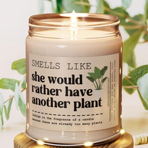 Könnte beinhalten: Eine beige Kerze in einem braunen Glasgefäß mit einem weißen Etikett, auf dem "Smells Like She Would Rather Have Another Plant" steht. Das Etikett enthält auch eine kleine Illustration einer Topfpflanze und den Text "Indulge in the fragrance of a candle because there are already too many plants".