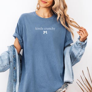 Op de afbeelding: Een blauw T-shirt met de tekst "kinda crunchy" en een witte strik bedrukt op de voorkant.