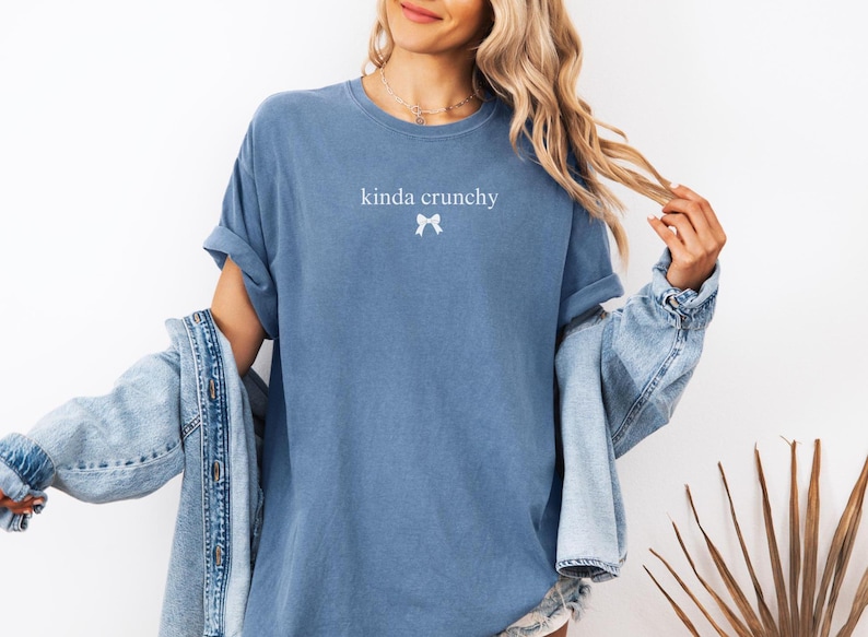 Op de afbeelding: Een blauw T-shirt met de tekst "kinda crunchy" en een witte strik bedrukt op de voorkant.