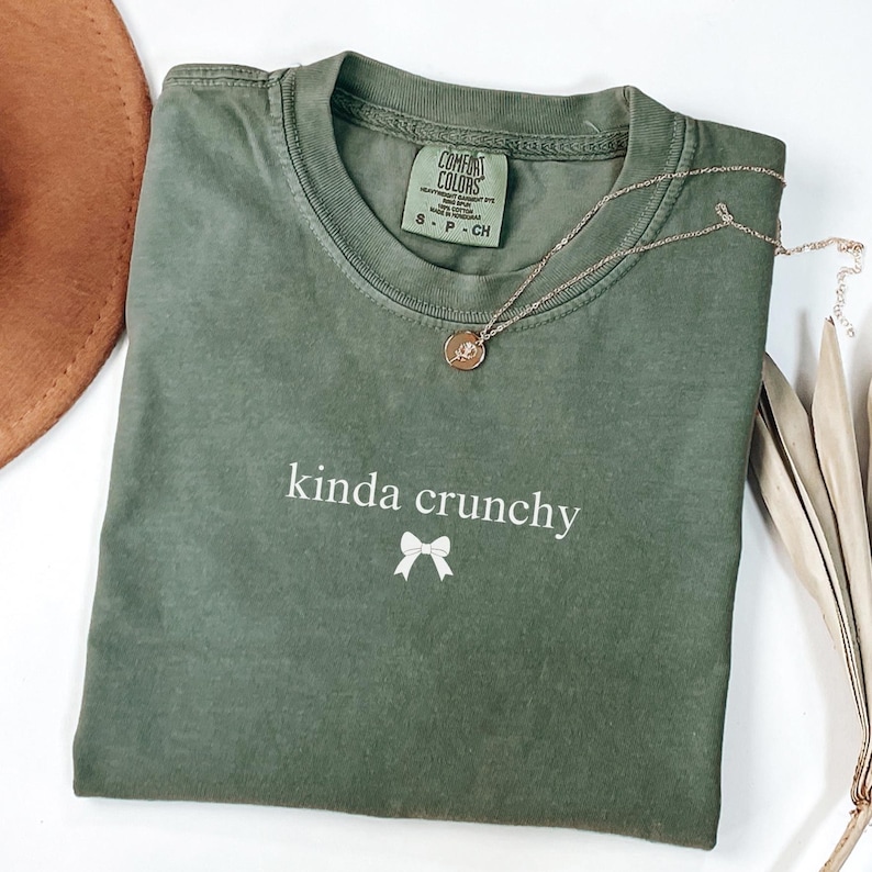 Op de afbeelding: Een groen T-shirt met de tekst "kinda crunchy" en een witte strik bedrukt op de voorkant.