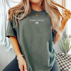 Op de afbeelding: Een donkergroen T-shirt met de tekst "kinda crunchy" en een witte strik bedrukt op de voorkant.