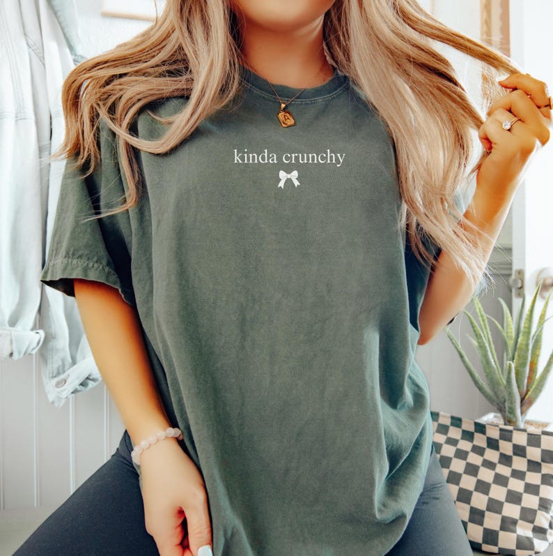 Op de afbeelding: Een donkergroen T-shirt met de tekst "kinda crunchy" en een witte strik bedrukt op de voorkant.