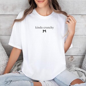 Op de afbeelding: Wit T-shirt met de tekst "kinda crunchy" en een klein zwart strikje.