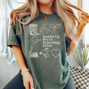 May include: A dark green t-shirt with white illustrations of cowboy boots, a cowboy on a horse, a rope, a cowboy hat, a cactus, a horse, and a heart. The text "Saddle Up We're Following Jesus" is printed in white.