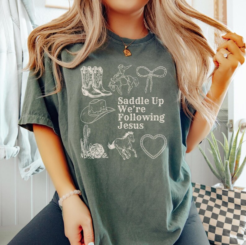 May include: A dark green t-shirt with white illustrations of cowboy boots, a cowboy on a horse, a rope, a cowboy hat, a cactus, a horse, and a heart. The text "Saddle Up We're Following Jesus" is printed in white.