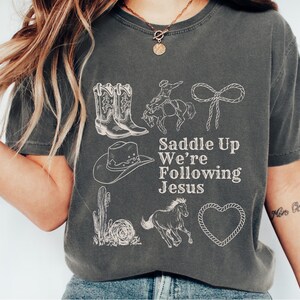 May include: Dark gray t-shirt with white illustrations of cowboy boots, a cowboy on a horse, a lasso, a cowboy hat, a cactus, a running horse, and a heart. The text "Saddle Up We're Following Jesus" is printed in white.