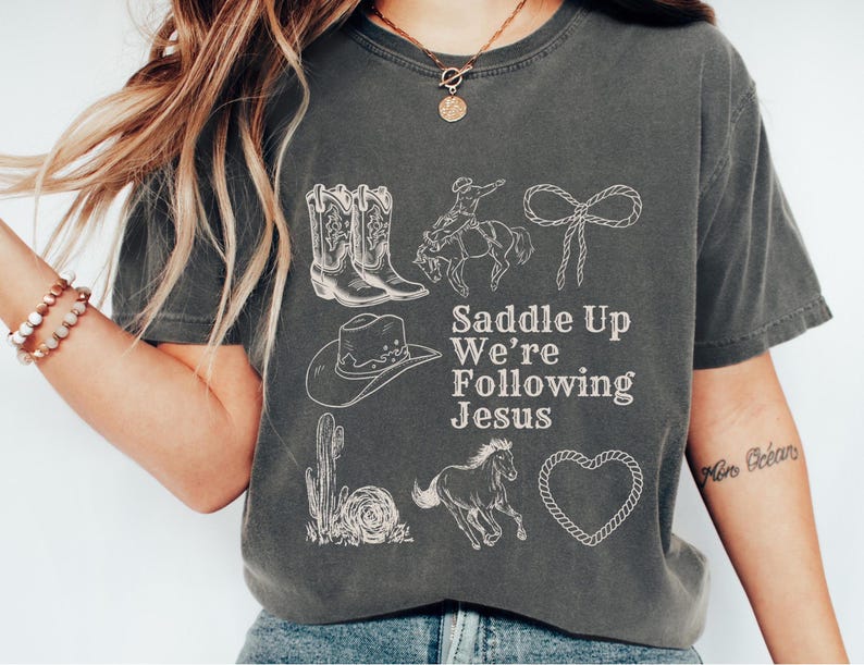 May include: Dark gray t-shirt with white illustrations of cowboy boots, a cowboy on a horse, a lasso, a cowboy hat, a cactus, a running horse, and a heart. The text "Saddle Up We're Following Jesus" is printed in white.