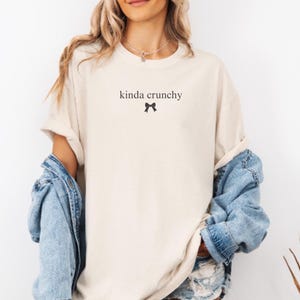 Op de afbeelding: Een vrouw draagt een lichtbeige t-shirt met de tekst "kinda crunchy" en een klein zwart strikje. Ze draagt een lichtblauw denim jack en een denim short.