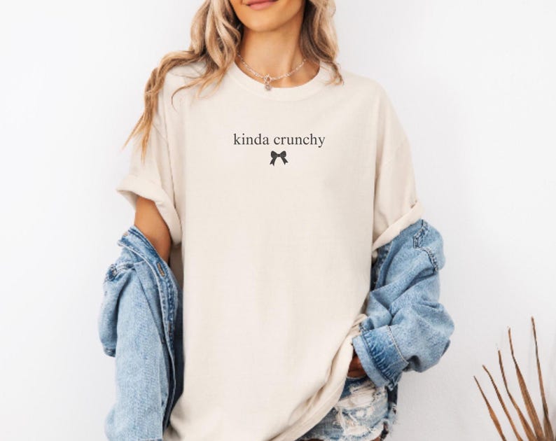 Op de afbeelding: Een vrouw draagt een lichtbeige t-shirt met de tekst "kinda crunchy" en een klein zwart strikje. Ze draagt een lichtblauw denim jack en een denim short.