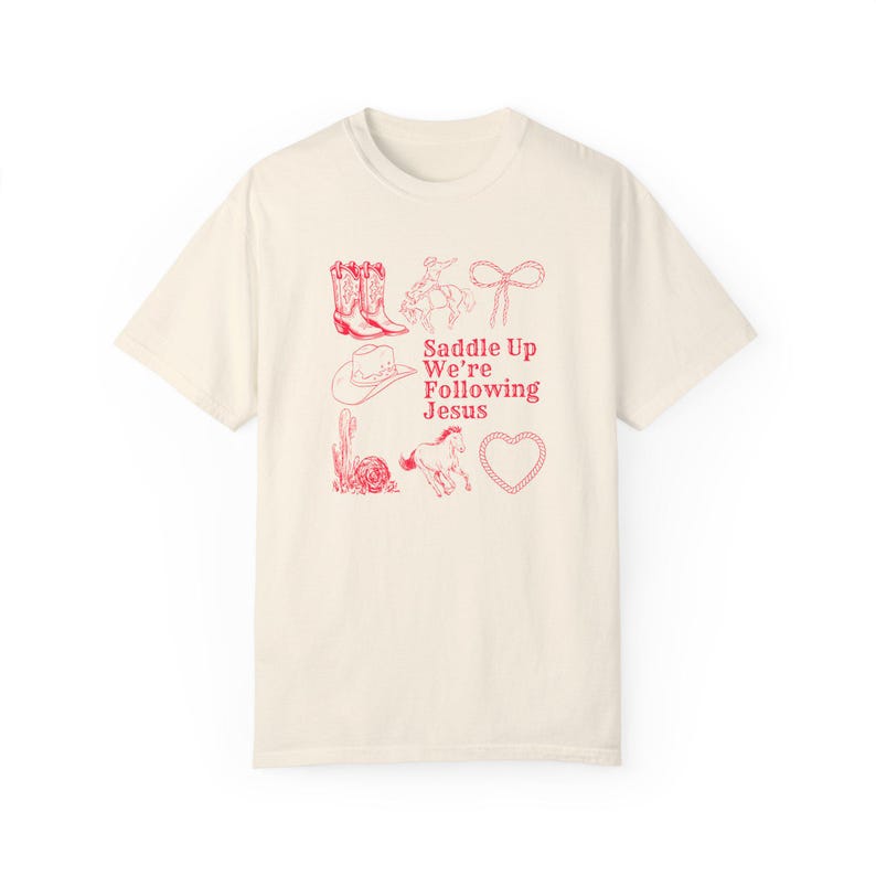 May include: Cream-colored t-shirt with a red graphic design featuring cowboy boots, a cowboy hat, a horse, a cactus, a lasso, and a heart. The text "Saddle Up We're Following Jesus" is also printed in red.
