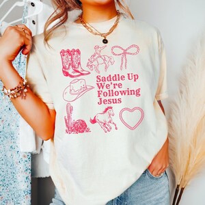 May include: Cream-colored t-shirt with a pink graphic design featuring cowboy boots, a cowboy on a horse, a cowboy hat, a cactus, a running horse, a heart, and the text "Saddle Up We're Following Jesus."