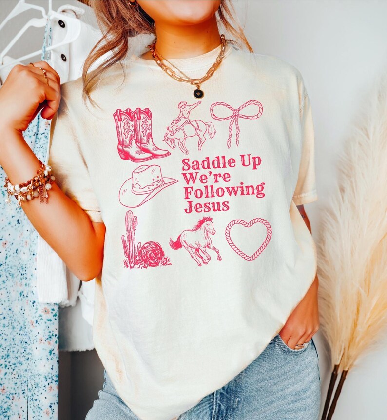 May include: Cream-colored t-shirt with a pink graphic design featuring cowboy boots, a cowboy on a horse, a cowboy hat, a cactus, a running horse, a heart, and the text "Saddle Up We're Following Jesus."