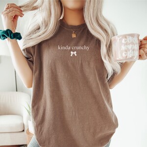 Op de afbeelding: Een oversized bruin t-shirt met de tekst "kinda crunchy" en een klein wit strikje geprint op de voorkant. Het model draagt het shirt met een groene scrunchie in haar haar.