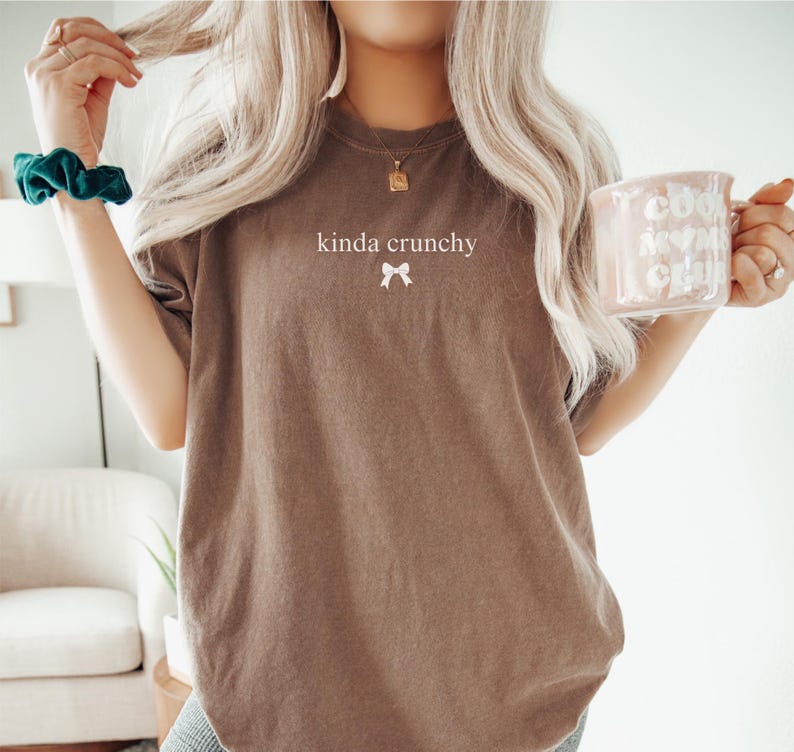 Op de afbeelding: Een oversized bruin t-shirt met de tekst "kinda crunchy" en een klein wit strikje geprint op de voorkant. Het model draagt het shirt met een groene scrunchie in haar haar.