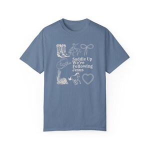 May include: A blue t-shirt with a white graphic design featuring cowboy boots, a cowboy on a horse, a lasso, a cowboy hat, a cactus, a horse, and a heart. The text "Saddle Up We're Following Jesus" is also included.