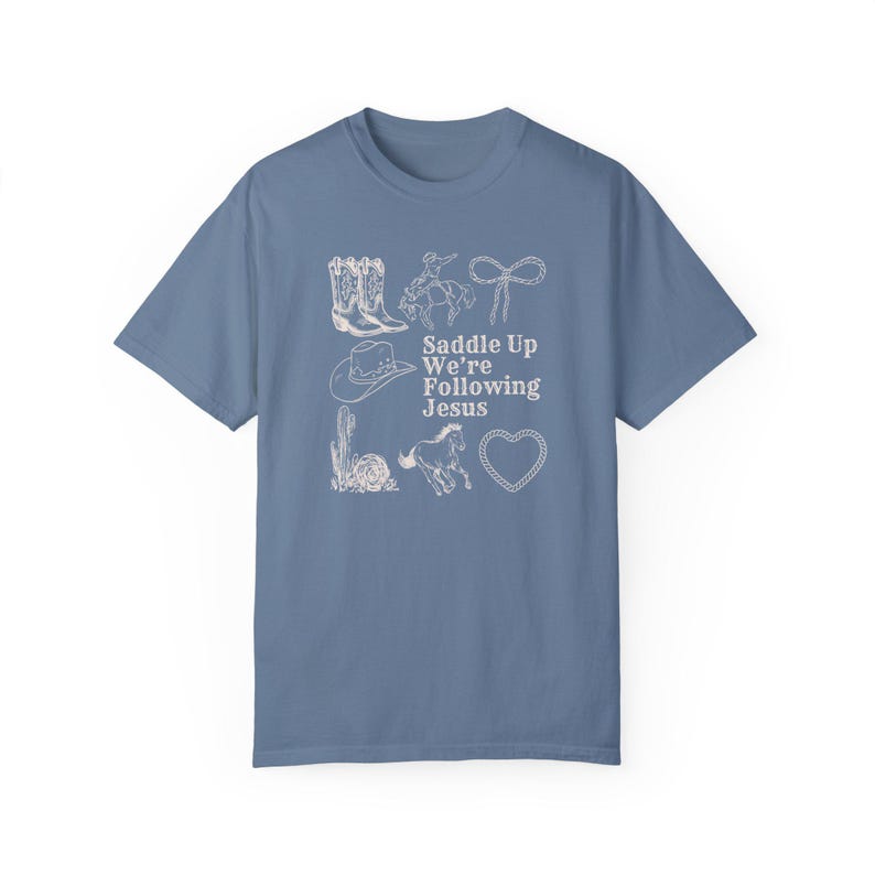 May include: A blue t-shirt with a white graphic design featuring cowboy boots, a cowboy on a horse, a lasso, a cowboy hat, a cactus, a horse, and a heart. The text "Saddle Up We're Following Jesus" is also included.