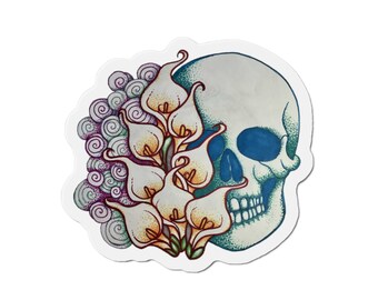 May include: A sticker featuring a detailed illustration of a skull with a blue eye socket and a bouquet of calla lilies. The flowers are white and yellow, and the design includes swirling purple and grey accents.