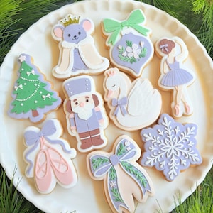 May include: A selection of decorated sugar cookies on a white plate. The cookies are shaped and decorated with icing to resemble a Christmas tree, a mouse king, a ballerina, a swan, a nutcracker, and a snowflake. The cookies are decorated with pastel colours and intricate details.