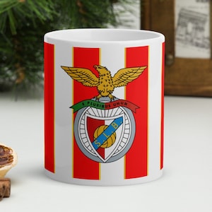 May include: White ceramic mug with a red and white striped design. The mug features a crest with a red shield, a green and white eagle, and the text "E PLURIBUS UNUM". The crest also includes a football and the letters "SLB".