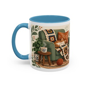 May include: A ceramic mug with a blue handle and interior featuring a printed illustration of a green armchair with a crocheted blanket, a sleeping orange tabby cat, a small wooden side table with a steaming cup, a potted plant, and yarn.