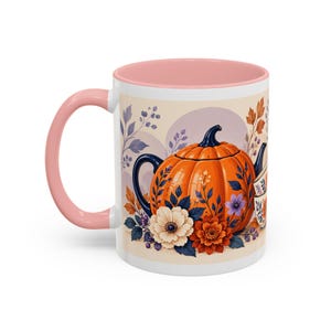 Pode incluir: Uma caneca de cerâmica com alça e borda rosa suave, apresentando uma ilustração gráfica de um bule laranja em forma de abóbora cercado por flores de outono coloridas, folhas e uma pilha de duas pequenas xícaras de chá em um fundo creme.