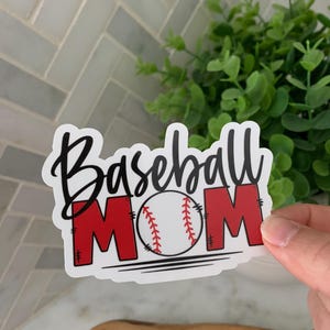 以下が含まれることがあります： 白地に黒文字で「Baseball Mom」と書かれたステッカー。中央には赤い野球ボールが描かれている。