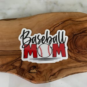 以下が含まれることがあります： 「Baseball Mom」という黒い文字と、中央に赤い野球のグラフィックが描かれた白いステッカーです。