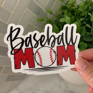 以下が含まれることがあります： 黒い文字で「Baseball Mom」と書いてある白いステッカー。スクリプトフォントを使用しています。「M」は赤で、中央には黒のステッチが入った白い野球ボールがあります。