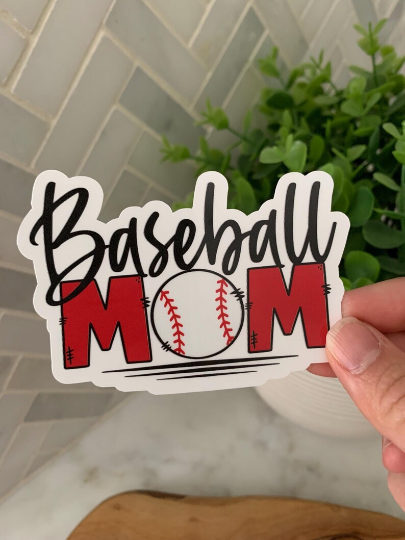 以下が含まれることがあります： 黒い文字で「Baseball Mom」と書いてある白いステッカー。スクリプトフォントを使用しています。「M」は赤で、中央には黒のステッチが入った白い野球ボールがあります。