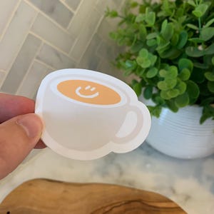 以下が含まれることがあります： カップの内側に笑顔が描かれた白いセラミック製のコーヒーカップ。カップは薄いオレンジ色の液体で満たされています。
