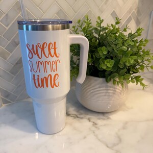 May include: A white insulated tumbler with a handle and a straw. The words "sweet summer time" are written in orange script on the front. The tumbler has a silver rim and a blue lid. A small potted plant is next to the tumbler.