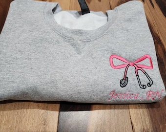 May include: Gray sweatshirt with a pink bow and a stethoscope embroidered on the right side. The text "Jessica, RN" is embroidered below the bow.