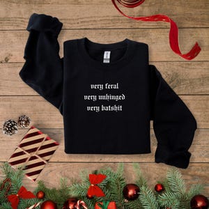 Puede incluir: Sudadera negra con texto blanco que dice "very feral very unhinged very batshit". La sudadera está sobre una superficie de madera con una cinta roja y ramas de pino.
