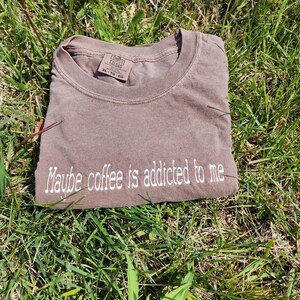 May include: A brown t-shirt with the text "Maybe coffee is addicted to me" embroidered in white on the front. The shirt is made by Comfort Colors and is size XL.