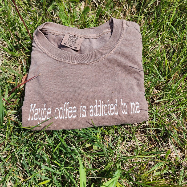 May include: A brown t-shirt with the text "Maybe coffee is addicted to me" embroidered in white on the front. The shirt is made by Comfort Colors and is size XL.