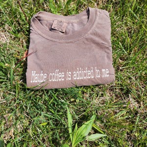 May include: A brown t-shirt with the text "Maybe coffee is addicted to me" embroidered in white on the front.