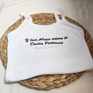 Könnte beinhalten: Weißes Tanktop mit dünnen Trägern und dem gestickten Text "If lost, please return to Dustin Patterson". Das Oberteil wird auf einer gewebten, kreisförmigen Matte präsentiert.