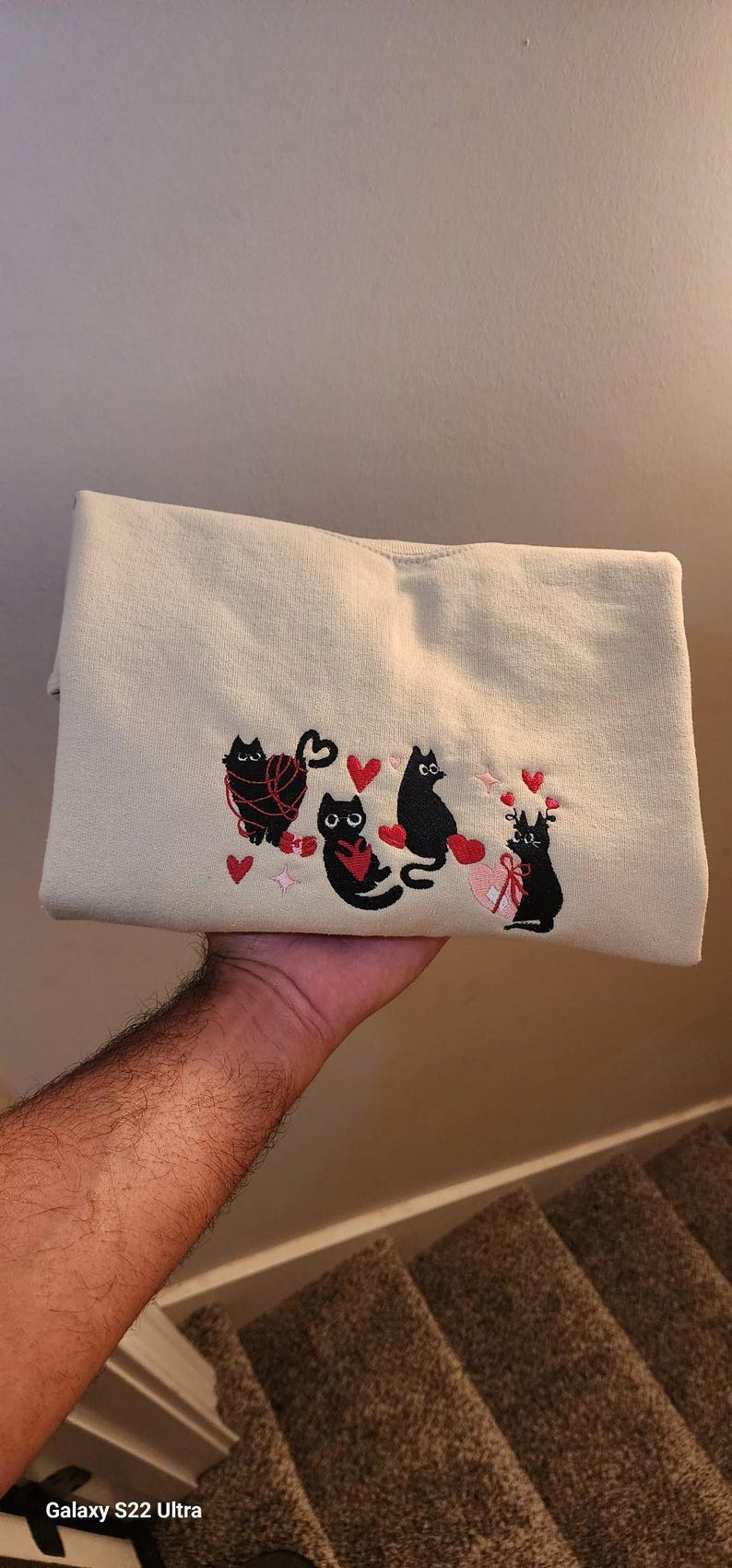May include: A cream-colored sweatshirt with black embroidered cats and red hearts. The cats are holding yarn balls and a gift box.