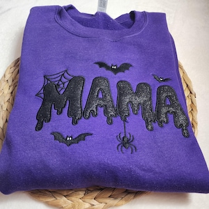 Könnte beinhalten: Ein violettes Sweatshirt mit Rundhalsausschnitt und dem Wort MAMA in einer schwarzen, glitzernden, tropfenden Schriftart. Das Design umfasst gestickte schwarze Fledermäuse, ein Spinnennetz in der oberen linken Ecke und eine hängende Spinne.