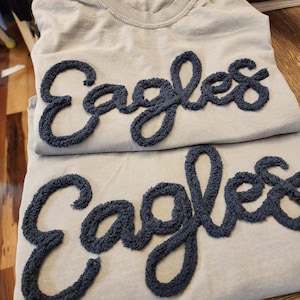 May include: Two light gray sweatshirts with the word "Eagles" embroidered in dark blue, textured lettering. The sweatshirts are folded and stacked on top of each other, showcasing the design.